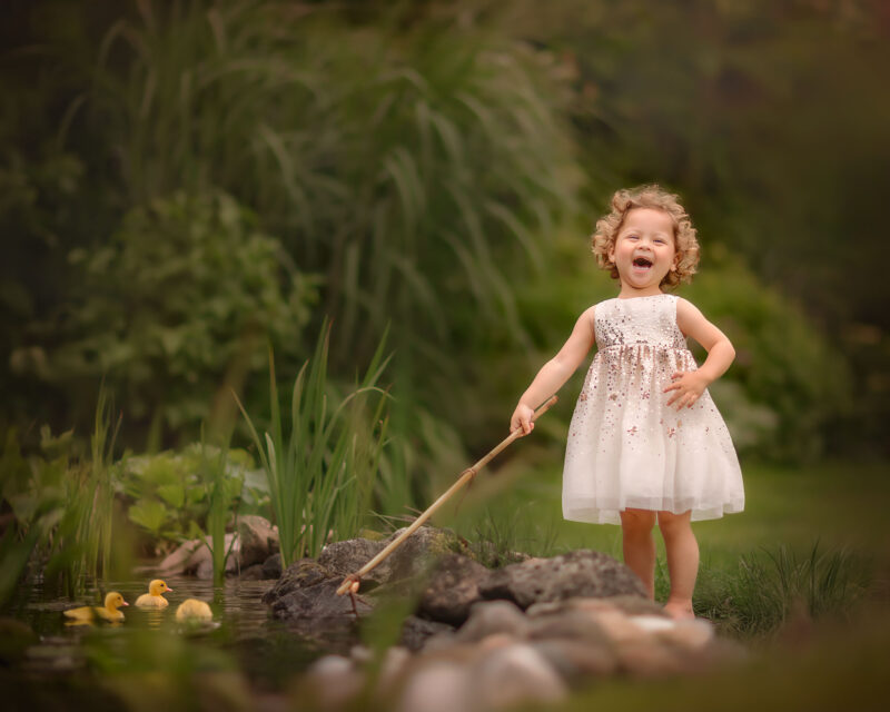 Fine-art-Canon-portret-van-een-lachend-klein-blond-krullend-meisje-bij-de-vijver-met-eendjes-door-natuurlijk-licht-fotograaf-Willie-Kers-uit-Apeldoorn